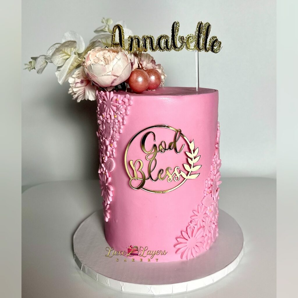 Elegant blush pink and white fondant christening cake with delicate floral appliqué accents, custom name topper, and God Bless plaque created by Luxe Layers Cakery in Miami Florida for a baby christening celebration.