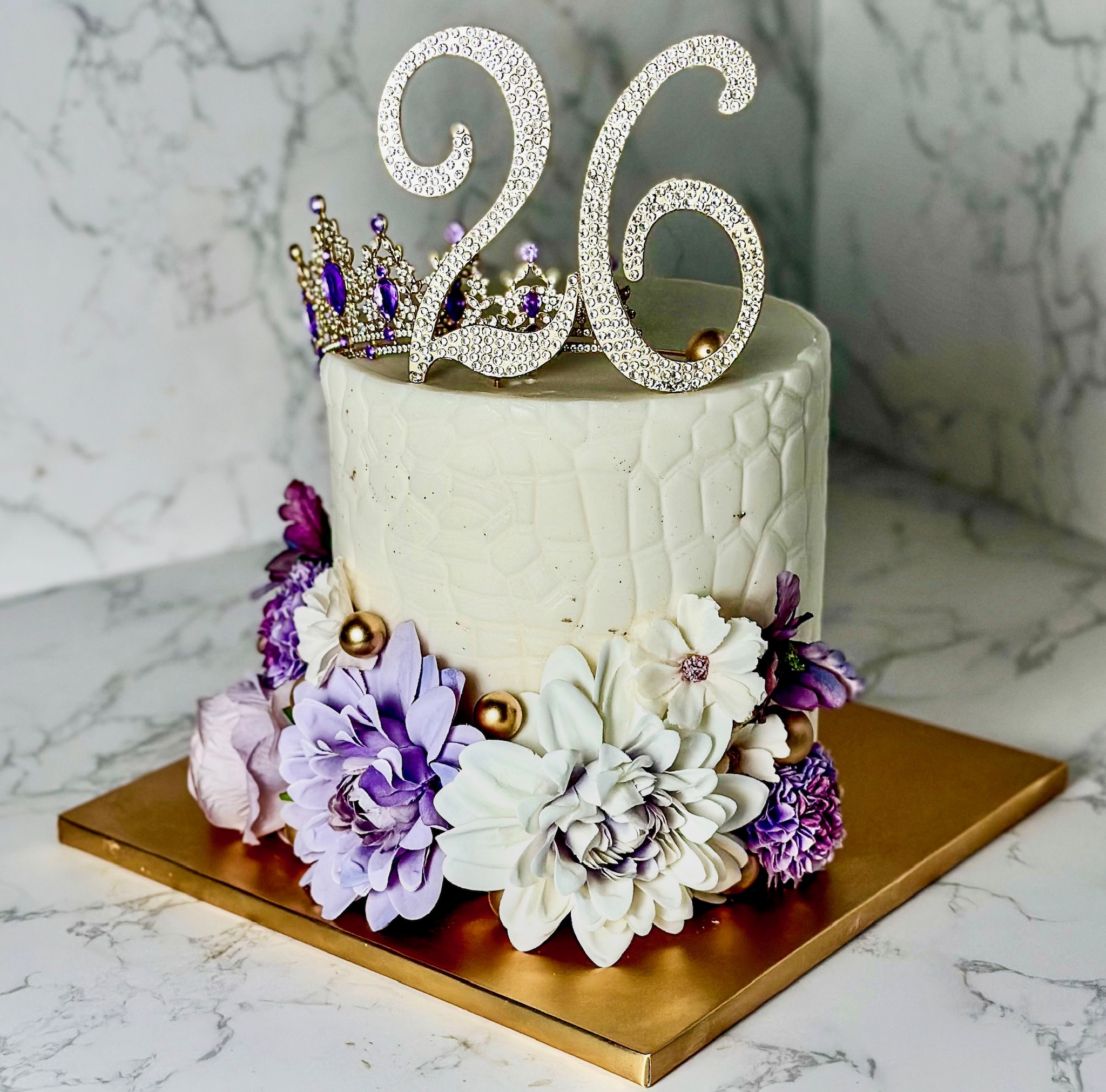 Single-tier white and lavender fondant 26th birthday cake with handcrafted floral arrangement, crown accent, gold spheres, and rhinestone topper — custom birthday cakes in Miami by Luxe Layers Cakery