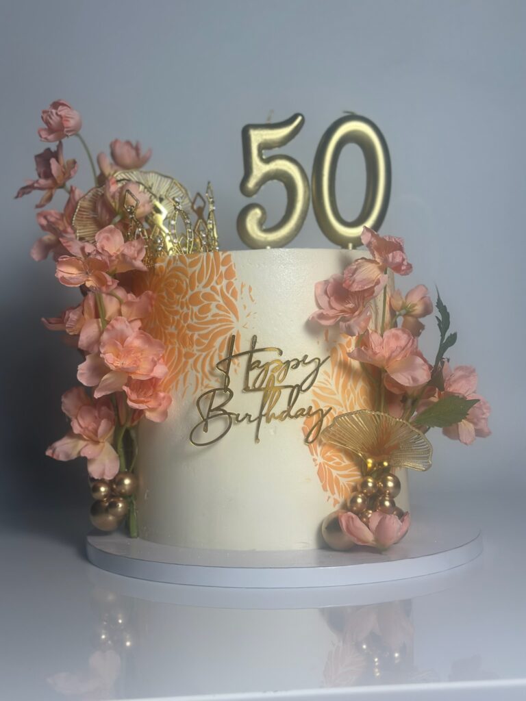 Single tier tropical pina colada 50th birthday cake with soft coral florals, gold metallic fan accents, decorative gold spheres, and Happy Birthday topper created by Luxe Layers Cakery in Miami Florida.