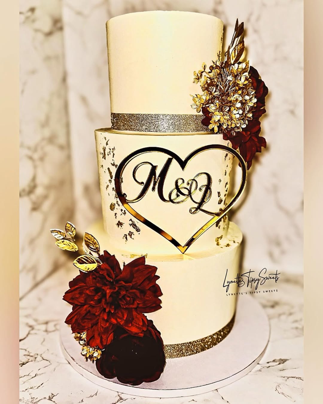 Three-tier ivory buttercream wedding cake with gold band detailing, gold leaf accents, deep burgundy florals, and custom heart-shaped acrylic monogram topper created by Luxe Layers Cakery in Miami Florida