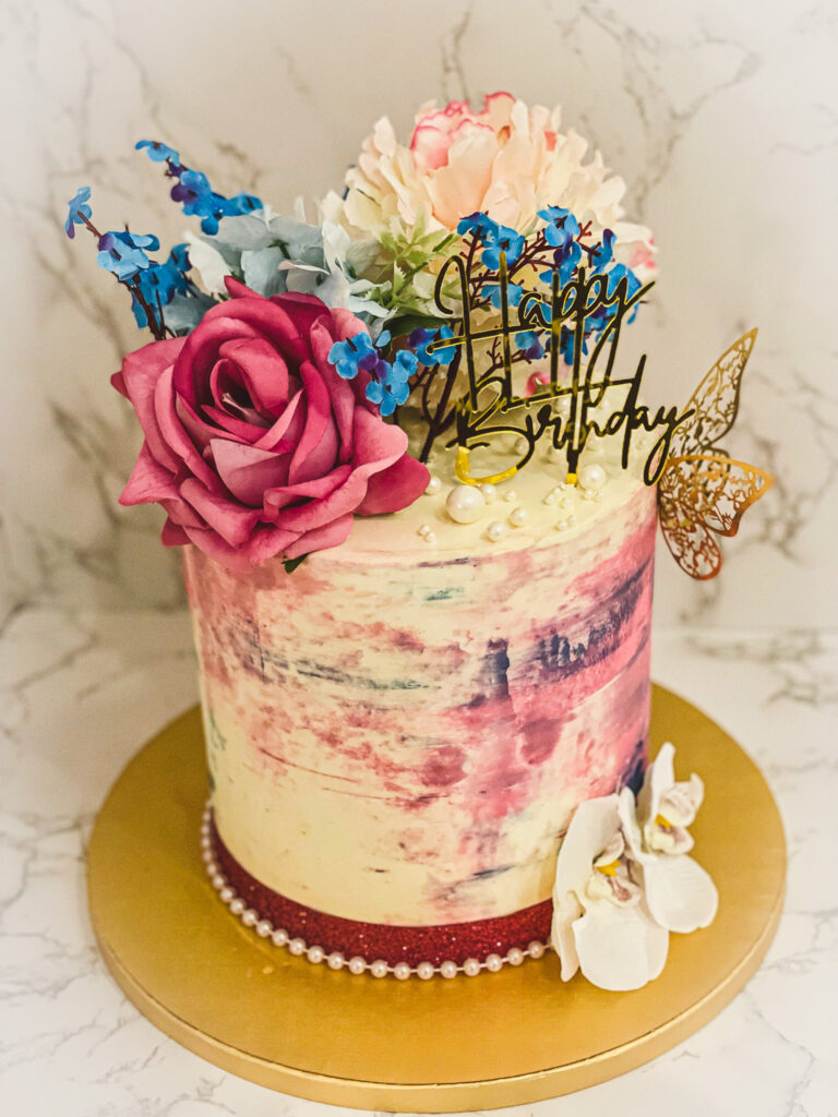 Modern floral painted buttercream birthday cake with soft pink and cream tones, gold Happy Birthday topper, decorative pearls, and butterfly accent, designed by Luxe Layers Cakery in Miami, Florida