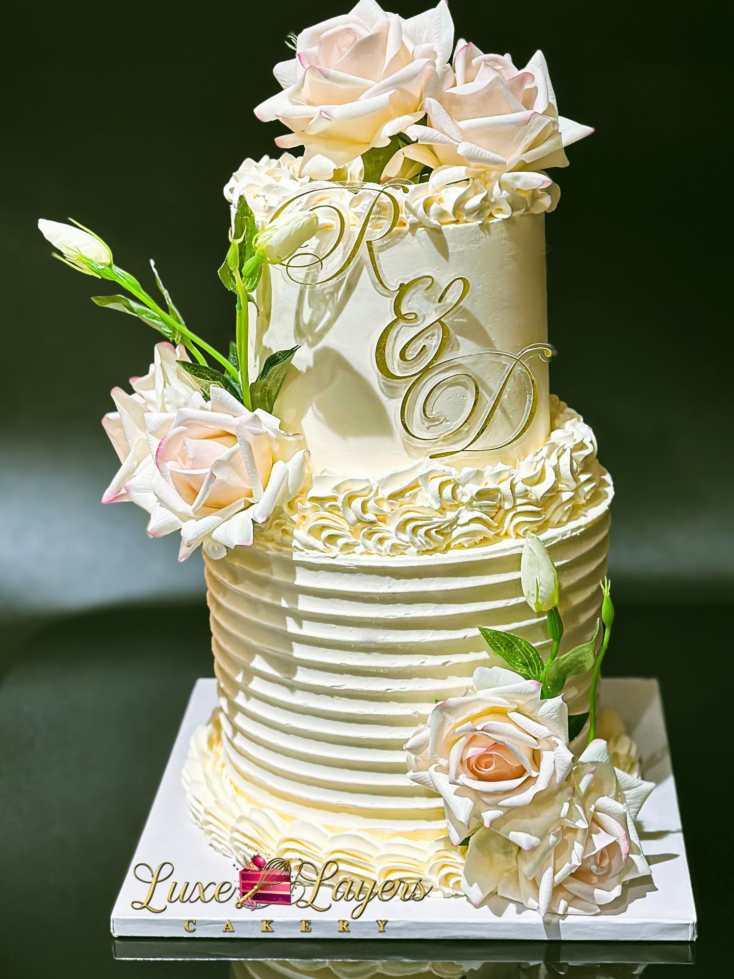 Two-tier garden romance wedding cake with smooth and horizontal textured buttercream finish, handcrafted ivory floral elements, piped border detailing, and custom gold monogram created by Luxe Layers Cakery in Miami Florida