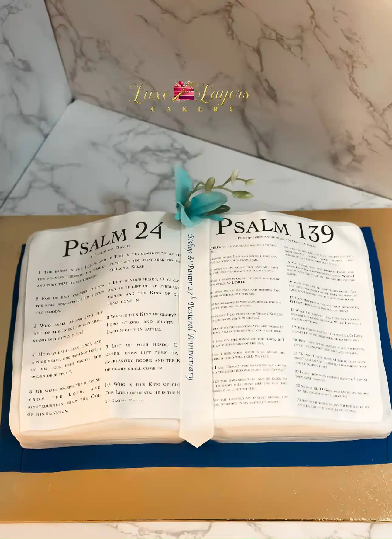 Open Bible-shaped cake with Psalm pages and blue floral accent, custom pastoral anniversary cake by Luxe Layers Cakery
