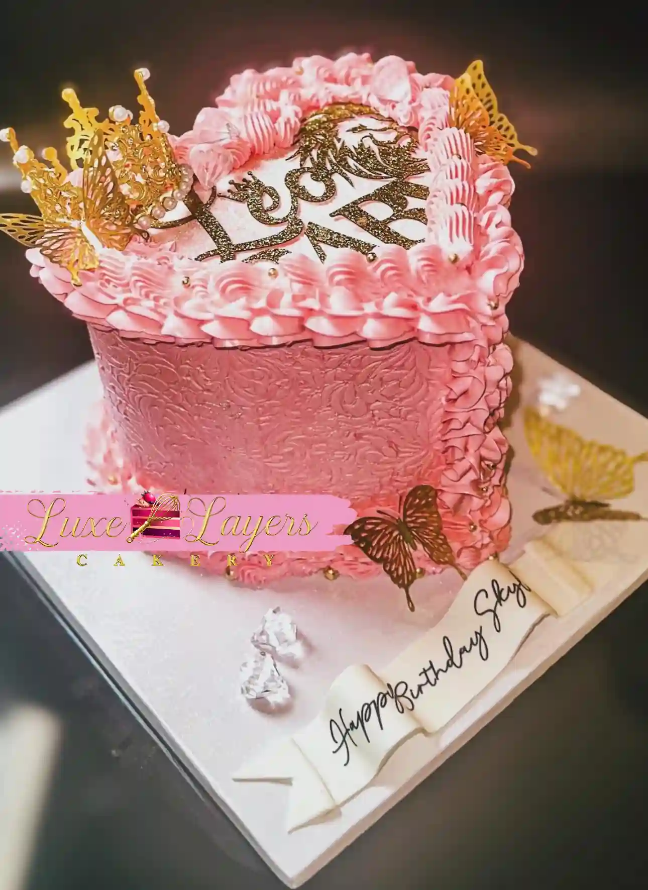Pink heart-shaped birthday cake with gold butterflies and crown topper, custom cake by Luxe Layers Cakery