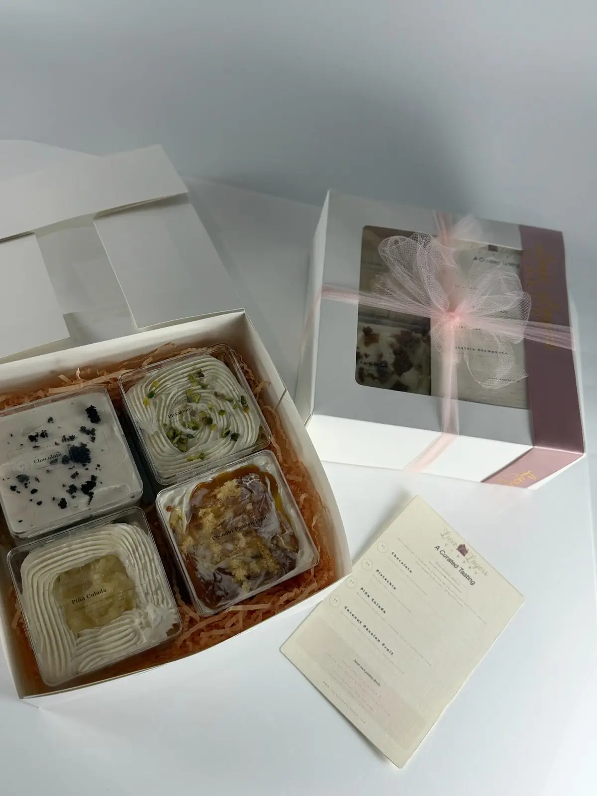 Open Luxe Layers Cakery cake taster box with four tasting containers beside a closed clear-window box and tasting menu
