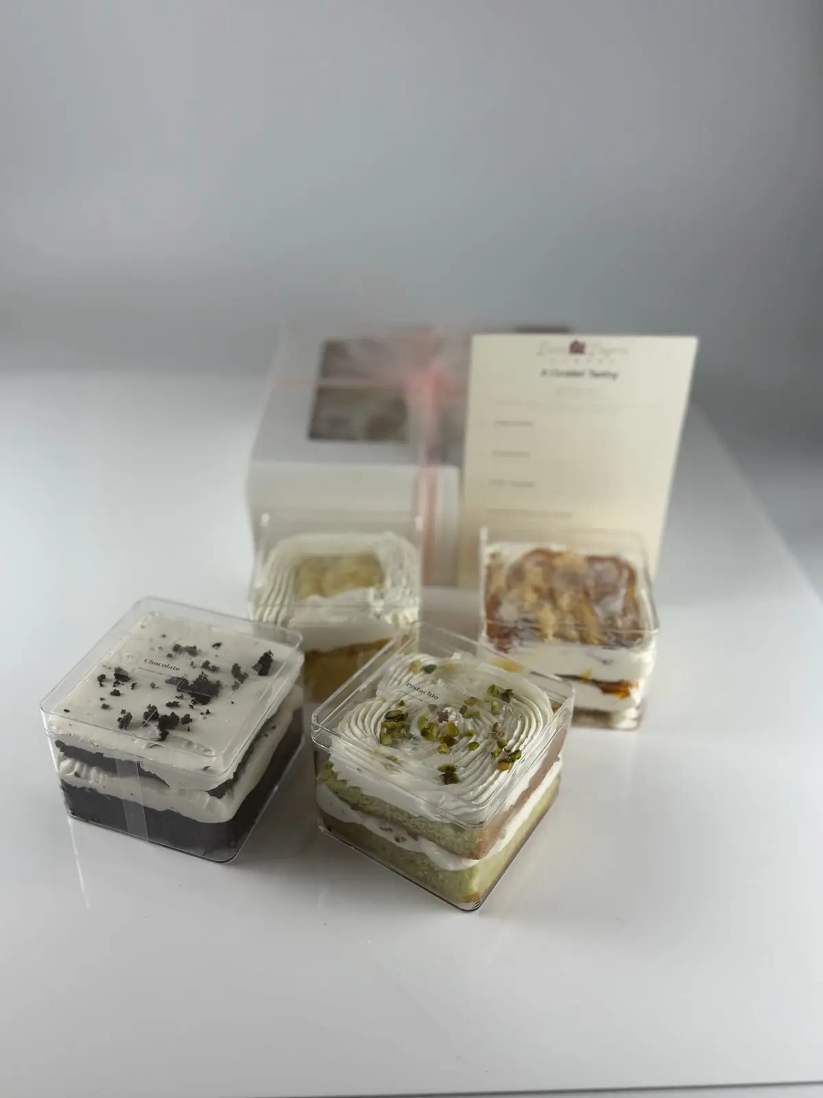 Four cake taster containers, including chocolate and pistachio, in front of a clear-window tasting box and menu card